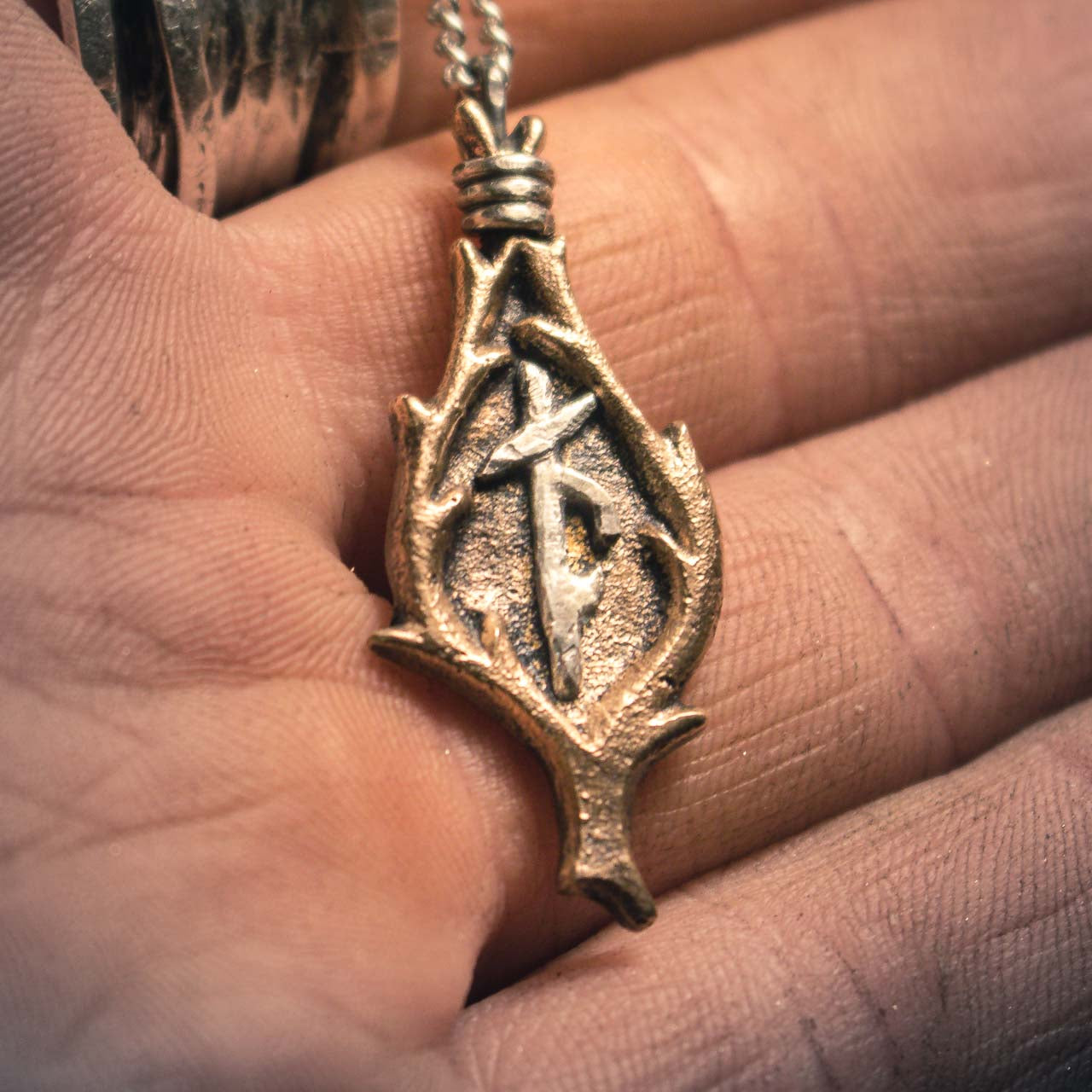 Close-up macro shot of a bronze Viking amulet with combined runes, suspended on a silver chain against a black surface
